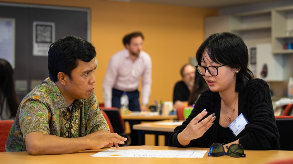 Two people engaged in discussion across a table during a workshop, with other participants in the background.