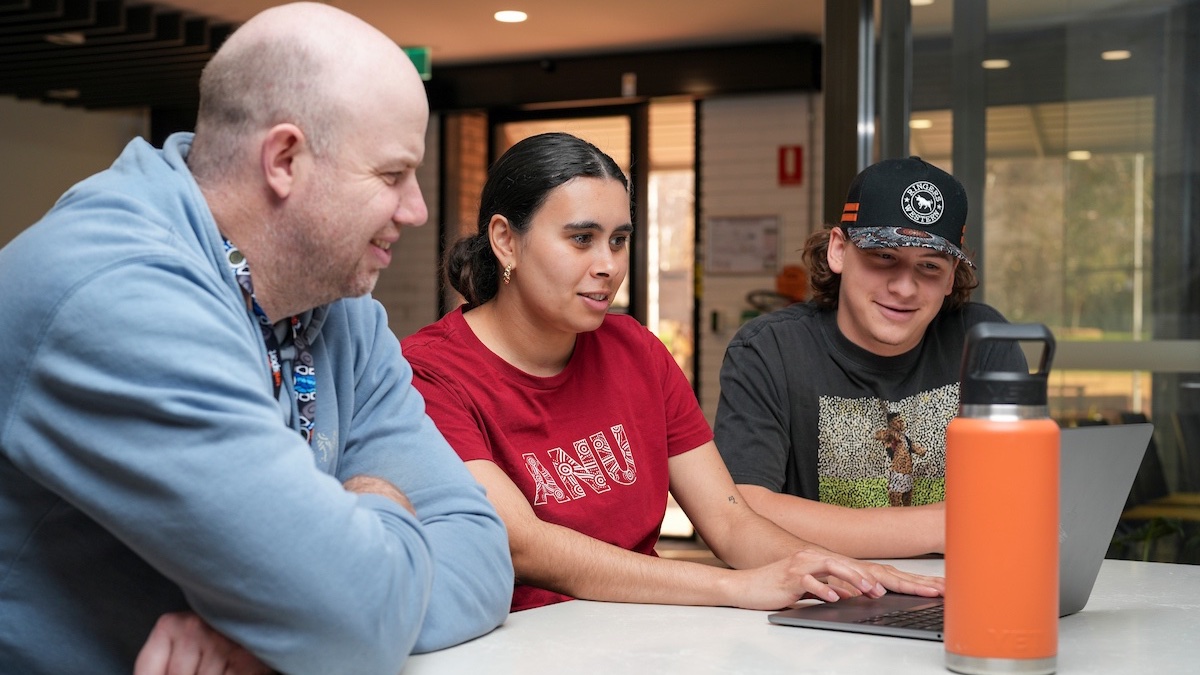 Three people sitting together looking at a laptop screen while in a discussion with each other. 